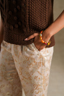 Close-up of a person wearing a chunky brown knit sweater and cream floral patterned trousers, with one hand in a pocket and a colorful beaded bracelet visible on the wrist.