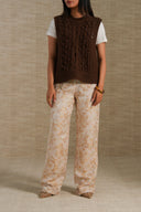 A model is shown from the neck down wearing a dark brown cable-knit sleeveless vest over a white t-shirt, paired with wide-leg beige trousers featuring a gold-toned floral pattern, set against a neutral textured beige backdrop.