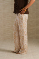 A person wearing wide-leg cream pants with a tan floral pattern, styled with a brown knit top and leopard-print shoes, standing against a textured beige backdrop.