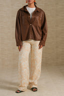 A model wearing a brown leather-look button-up jacket over a white t-shirt, paired with beige and orange patterned wide-leg trousers and black slide sandals, standing against a textured beige backdrop.